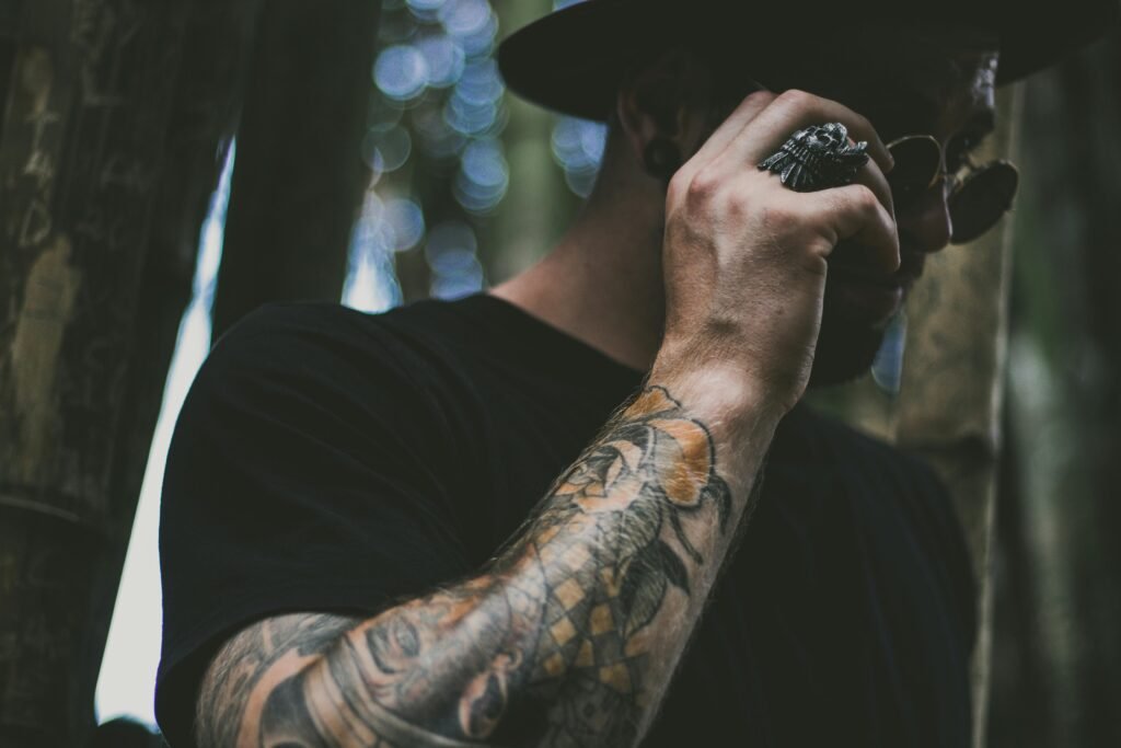 Tattooed man adjusting sunglasses amidst bokeh effect in forest setting.